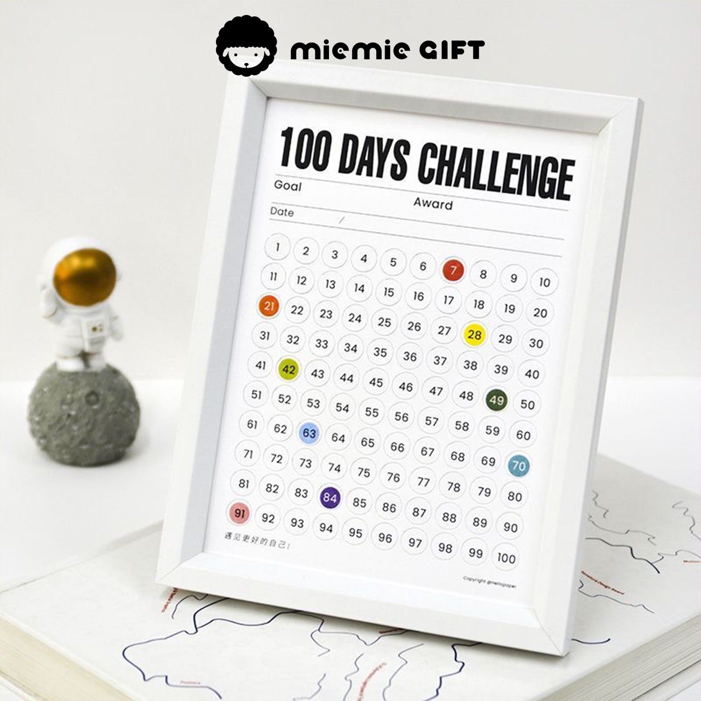 A white-framed 100 Days Challenge tracker placed on a desk, featuring numbered circles to mark daily progress. The frame is displayed alongside an astronaut figure, emphasizing motivation and creativity. Designed by MieMie Gift.