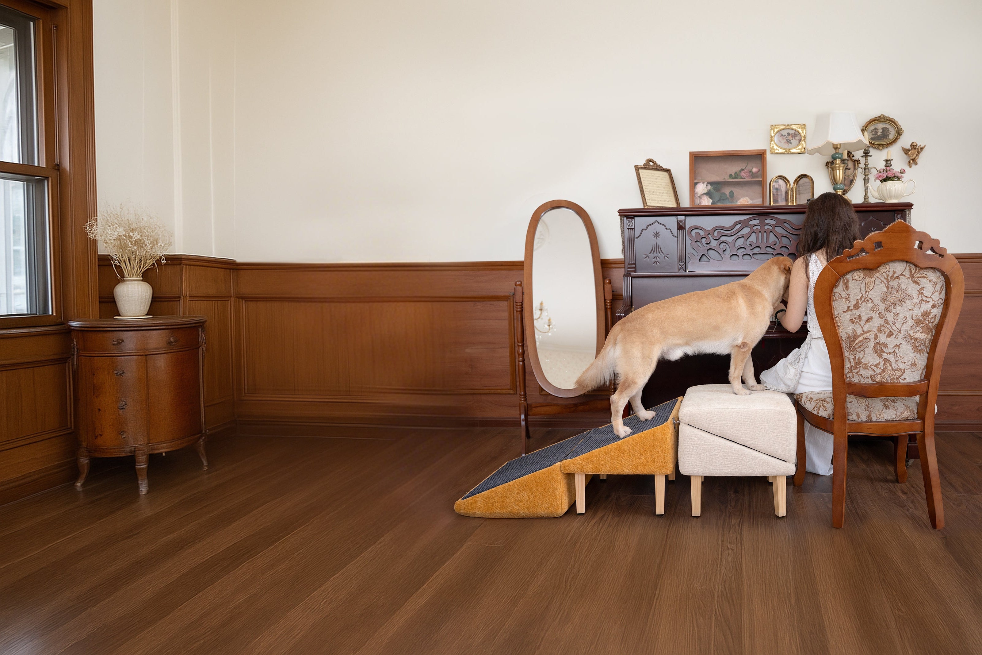 A golden retriever climbing the MieMie’s Corner dog ramp and pet stairs, designed to assist pets with joint issues or mobility limitations. The multi-level ramp provides a safe and comfortable way for dogs to reach elevated surfaces like sofas or beds. Set in a warm living room with wooden floors and vintage furniture, the dog steps onto a cushioned ottoman beside a woman seated at a piano.