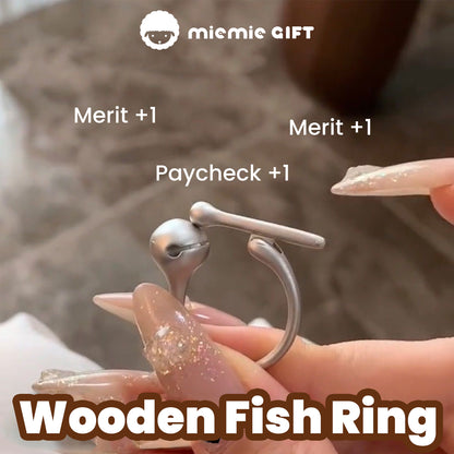 A close-up of the Wooden Fish Silver Ring being tapped, with text reading 'Merit +1, Paycheck +1,' highlighting its playful and stress-relief design in 925 sterling silver, by MieMie Gift.