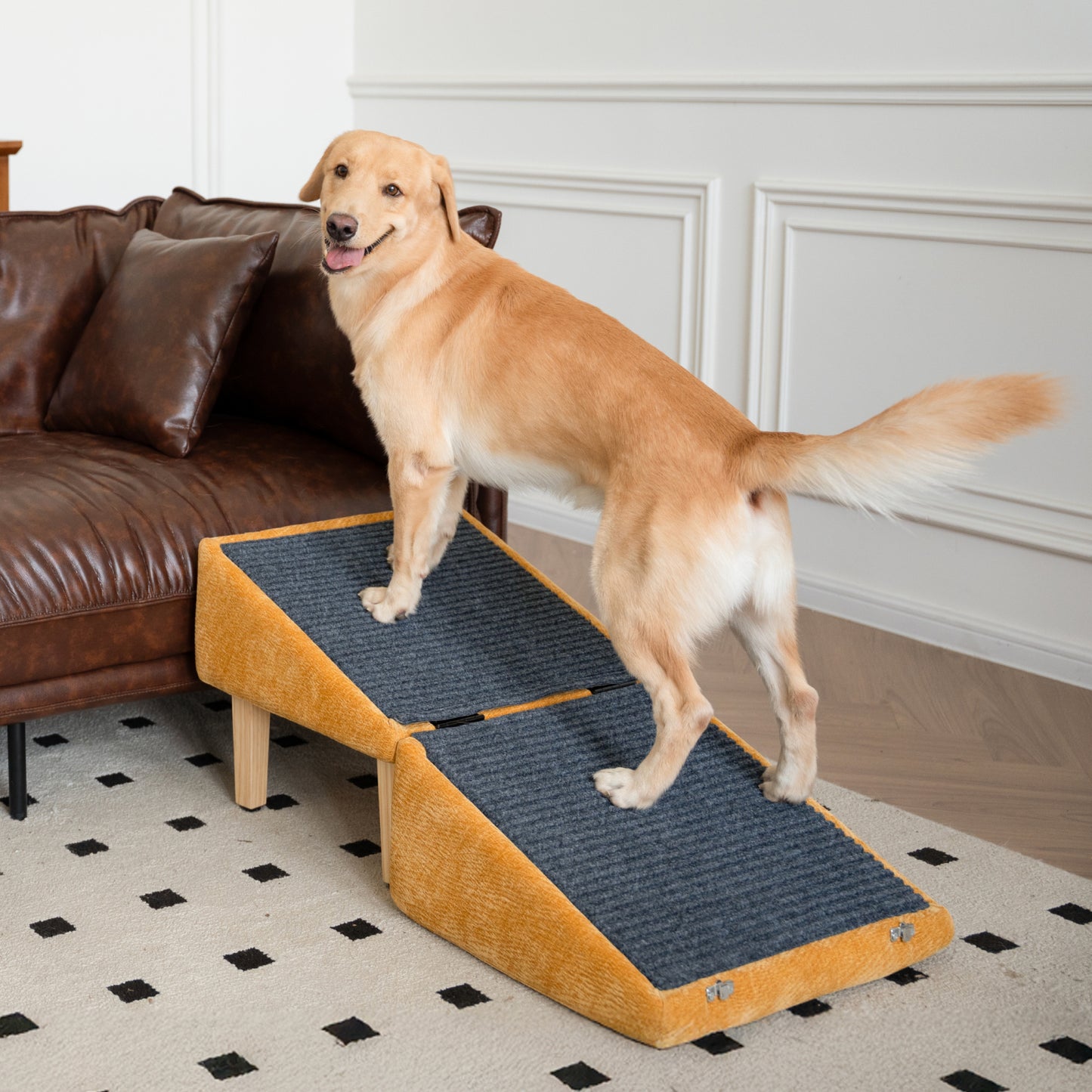 A cheerful golden retriever standing confidently on a mustard-yellow dog ramp in a modern living room. The ramp leads up to a brown leather sofa and features a foldable, non-slip carpeted surface with wooden support legs. The dog is looking back playfully at the camera, tail wagging, showing comfort and trust in using the ramp. The room is styled with a black-and-white patterned rug and white wall paneling.
